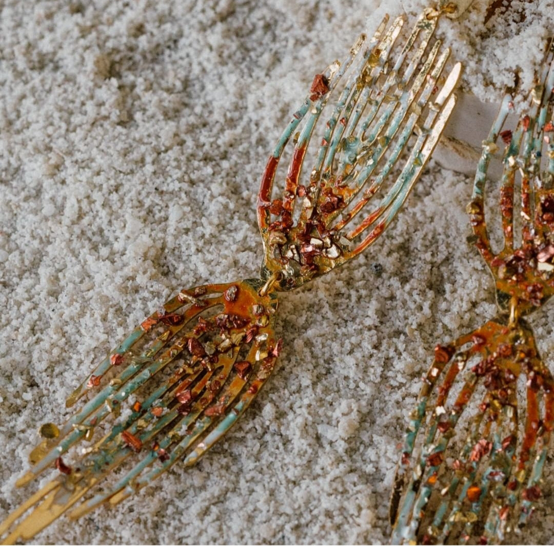 Pair of handcrafted gold earrings shaped like hands, decorated with orange, red, and turquoise crystal fragments, placed on sand