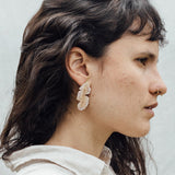 Portrait of a woman wearing the Banana Leaf Earrings—hand-painted sage-green and rust variants with glass granulation in gold-plated bronze—posed against a backdrop of large green leaves.