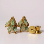 Flat lay of two Topos Nebula Mini leaf-shaped stud earrings hand-painted in green with glass granulation, accompanied by a separate round gold-plated earring back on a white surface