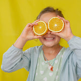 “Child wearing a light denim jacket and mint top holds two oranges over her eyes against a yellow backdrop.