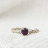 Close-up of a sterling silver ring featuring a 4mm round amethyst set in a textured band, displayed on soft white fabric.