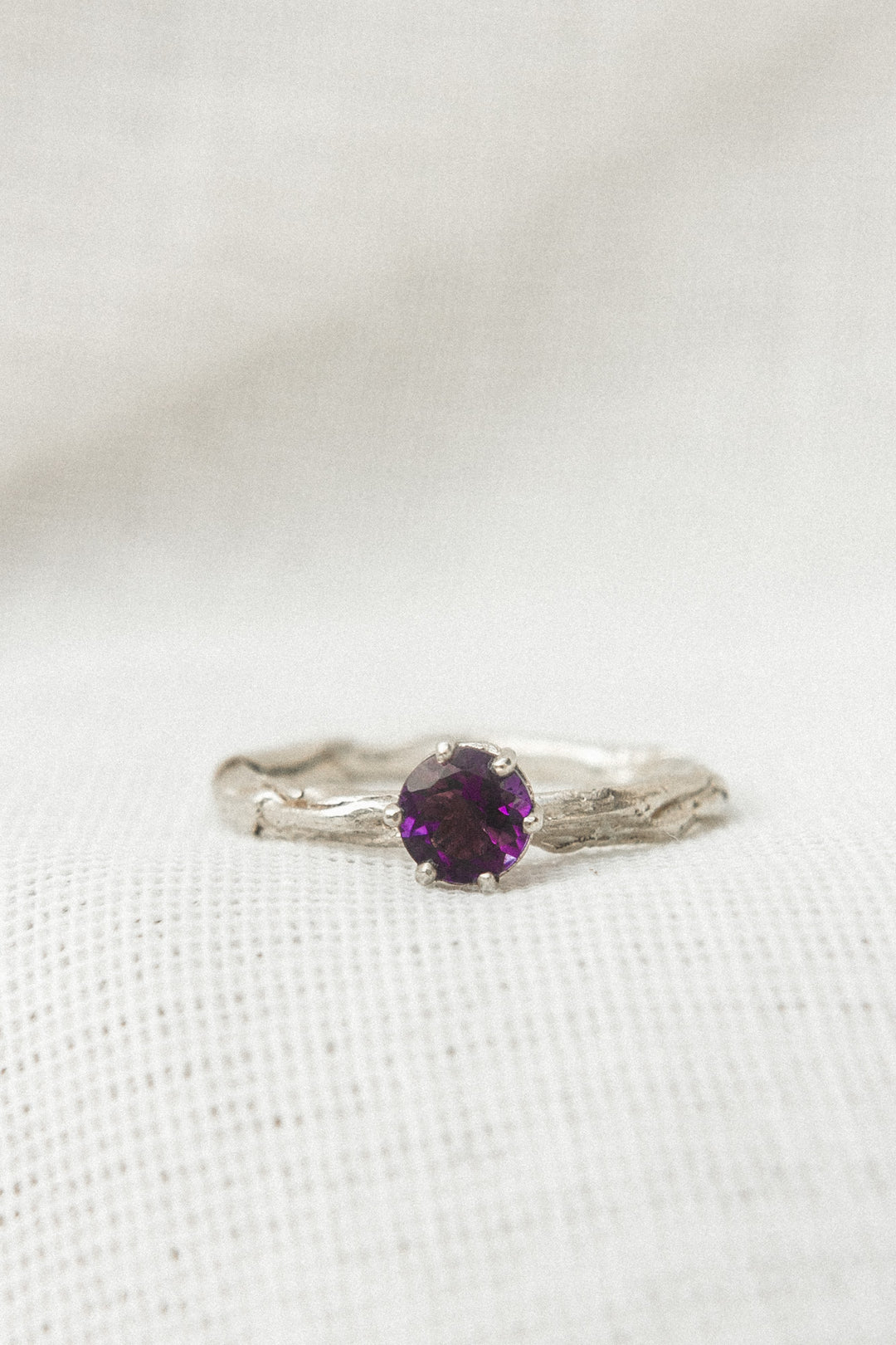 Close-up of a sterling silver ring featuring a 4mm round amethyst set in a textured band, displayed on soft white fabric.