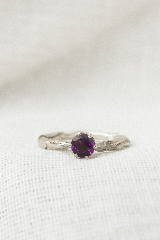 Close-up of a sterling silver ring featuring a 4mm round amethyst set in a textured band, displayed on soft white fabric.