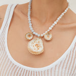 Model wearing a statement necklace with three gold and white shell-shaped pendants on a beaded white and light blue chain.