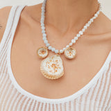 Model wearing a statement necklace with three gold and white shell-shaped pendants on a beaded white and light blue chain.