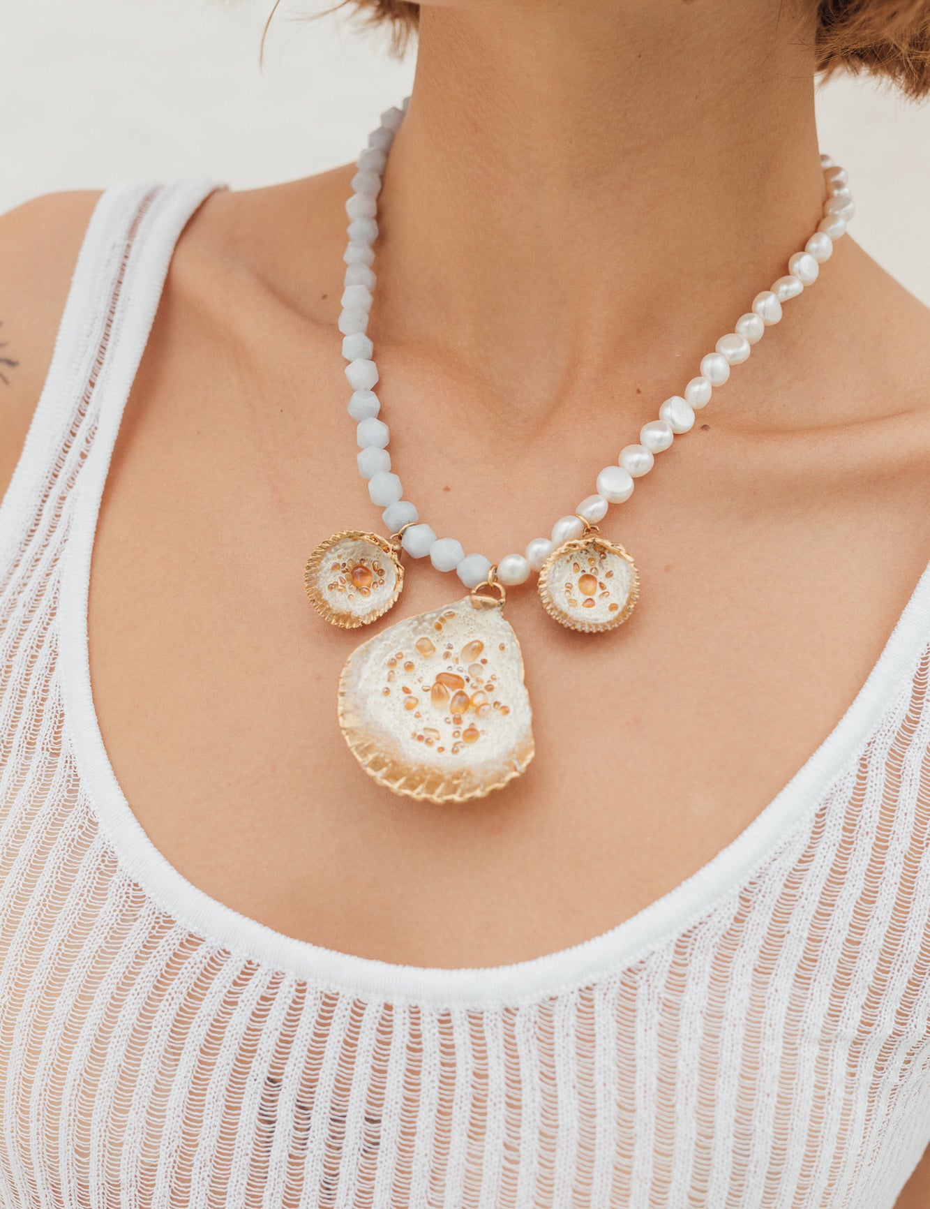 Model wearing a statement necklace with three gold and white shell-shaped pendants on a beaded white and light blue chain.