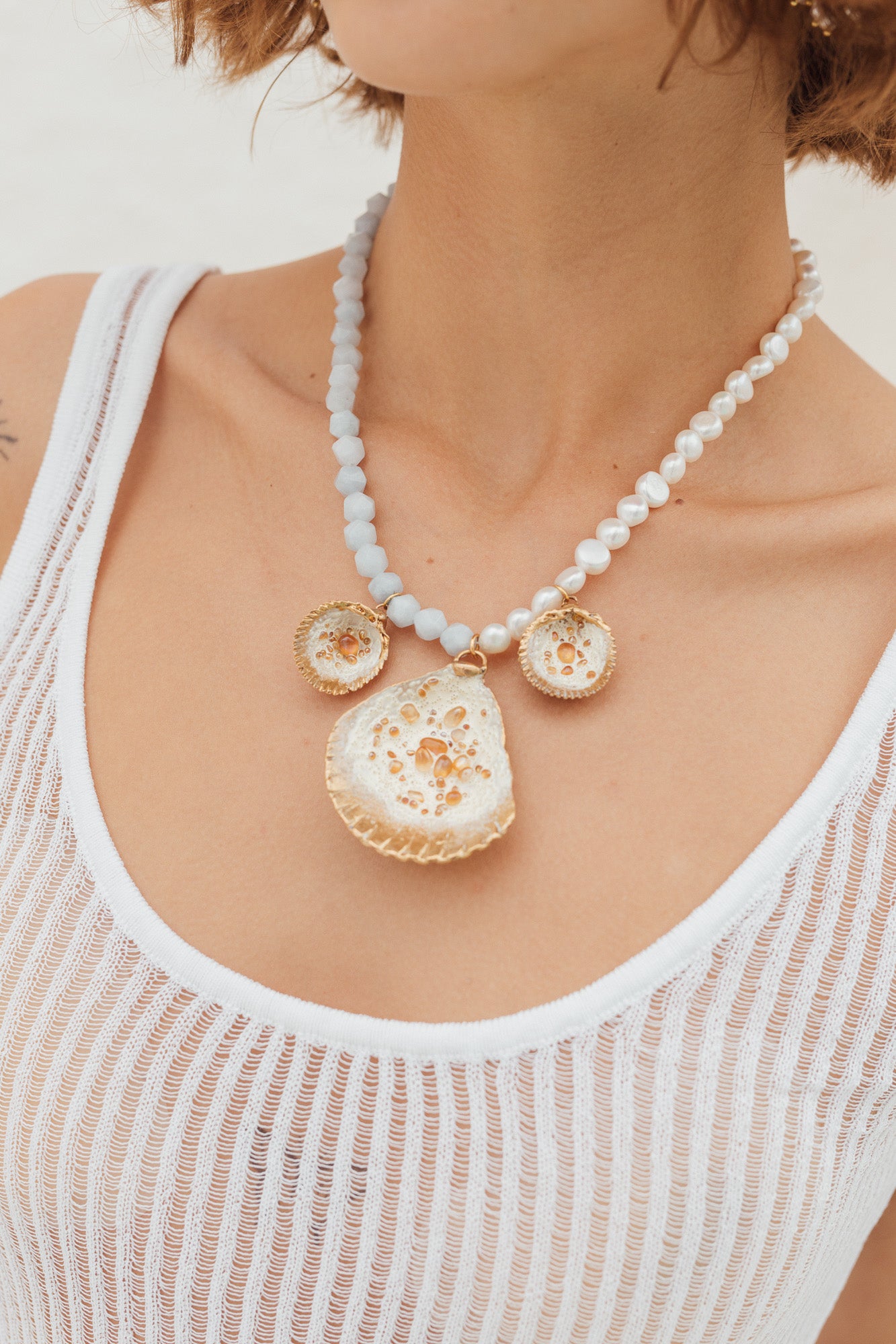 Model wearing a statement necklace with three gold and white shell-shaped pendants on a beaded white and light blue chain.