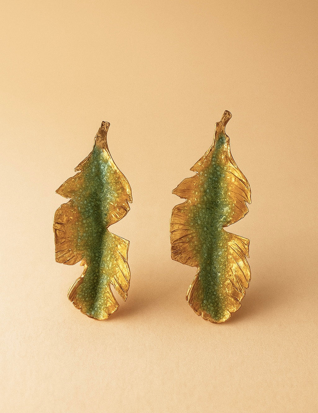 Pair of Banana Leaf Earrings in gold-plated brass hand-painted sage-green with glass granulation detail, displayed against a soft peach background.