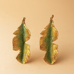 Pair of Banana Leaf Earrings in gold-plated brass hand-painted sage-green with glass granulation detail, displayed against a soft peach background.