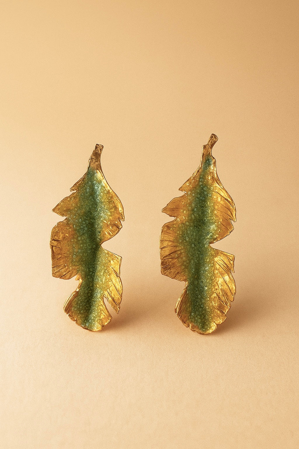 Pair of Banana Leaf Earrings in gold-plated brass hand-painted sage-green with glass granulation detail, displayed against a soft peach background.