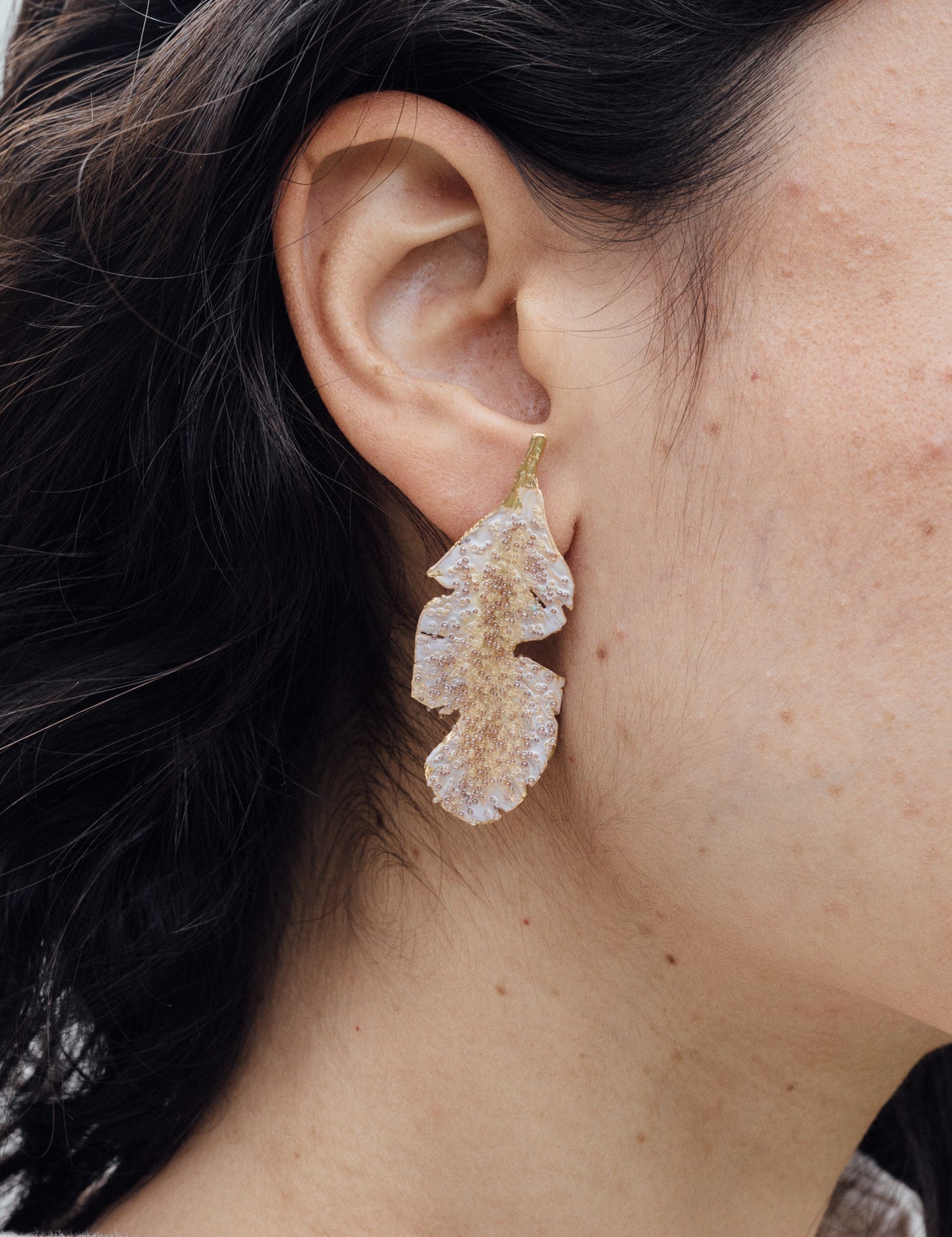 “Close-up side view of a woman’s ear wearing a single Banana Leaf Earring painted in sage-green with glass granulation on a gold-plated bronze setting, hair tucked behind ear against a neutral background.