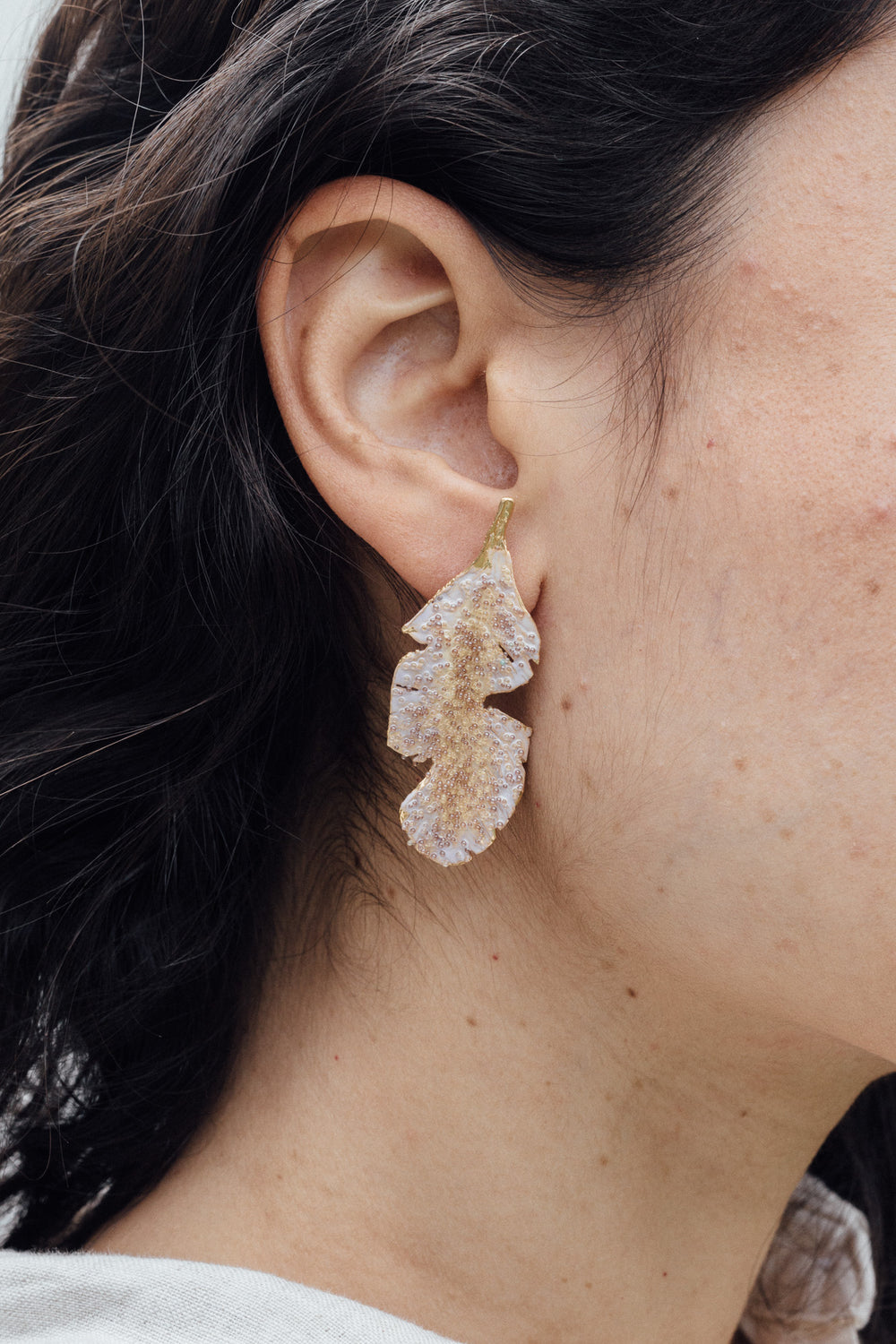 “Close-up side view of a woman’s ear wearing a single Banana Leaf Earring painted in sage-green with glass granulation on a gold-plated bronze setting, hair tucked behind ear against a neutral background.