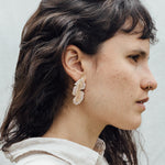 Portrait of a woman wearing the Banana Leaf Earrings—hand-painted sage-green and rust variants with glass granulation in gold-plated bronze—posed against a backdrop of large green leaves.