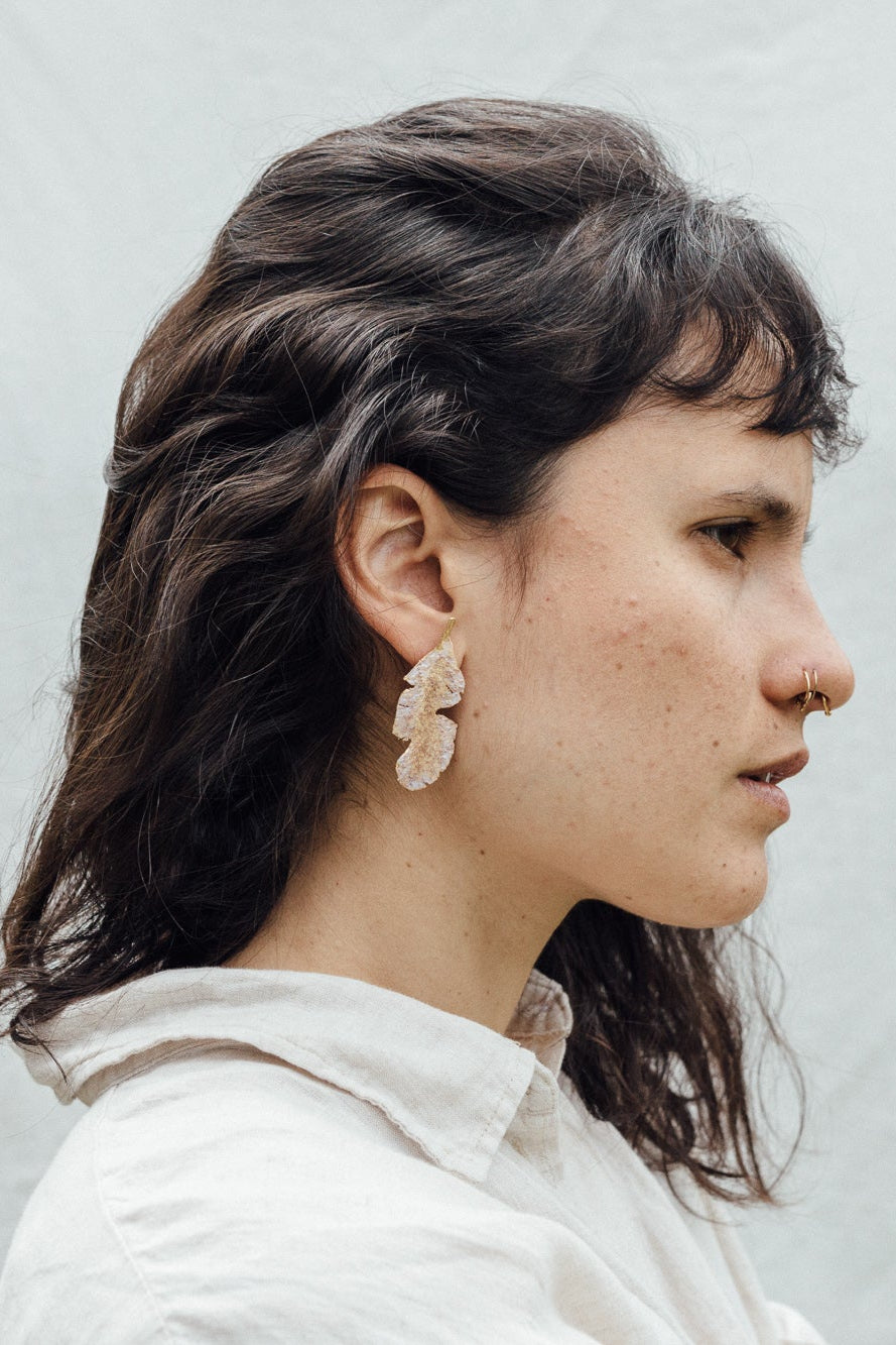 Portrait of a woman wearing the Banana Leaf Earrings—hand-painted sage-green and rust variants with glass granulation in gold-plated bronze—posed against a backdrop of large green leaves.