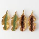 Flat lay of four Banana Leaf Earrings arranged horizontally on a white background—two hand-painted in sage-green and two in rust—with glass granulation and gold-plated bronze settings.