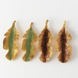 Flat lay of four Banana Leaf Earrings arranged horizontally on a white background—two hand-painted in sage-green and two in rust—with glass granulation and gold-plated bronze settings.