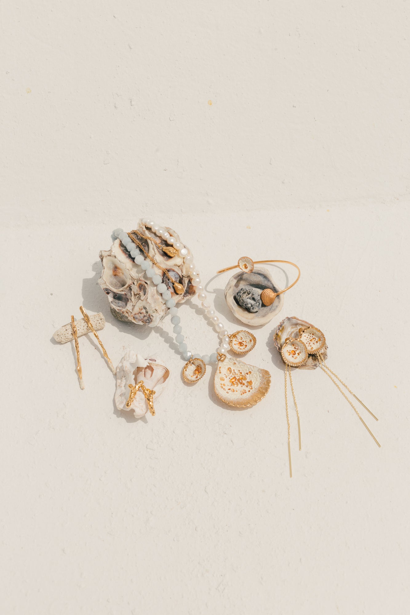 Flat lay of ocean-inspired jewelry, including gold-dipped shells, pearl and gemstone necklaces, earrings, and hairpins, displayed on a white textured surface.