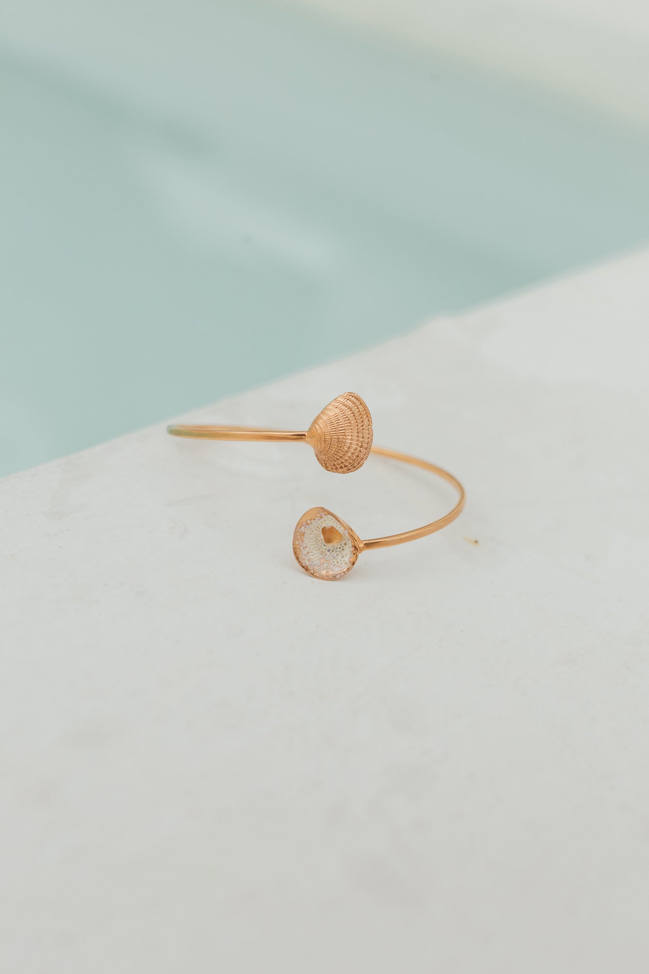 Delicate gold bracelet with open ends featuring a detailed shell and a resin-filled seashell, displayed beside a pool