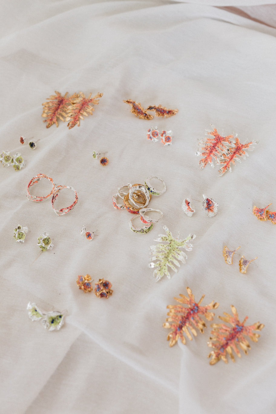 Colorful assortment of handcrafted rings and earrings inspired by nature, featuring floral and coral-like designs in gold, silver, and vibrant resins, displayed on a soft white fabric.