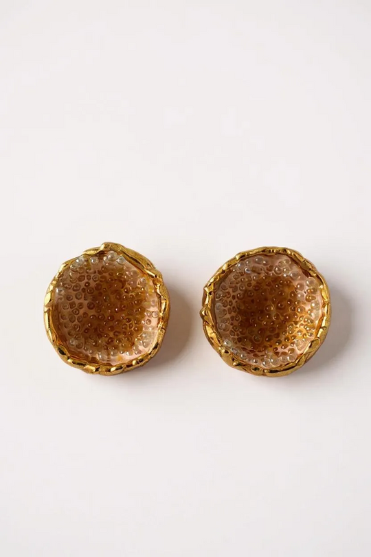 Flat lay of a pair of Topos Coca M stud earrings in gold-plated brass, hand-painted in sunset with turquoise glass granulation, on a white background.
