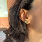 Close-up side view of a woman’s ear wearing a  Coca M stud earring, hand-painted in sage with glass granulation on a gold-plated brass setting, her hair tucked behind her ear against a neutral background.