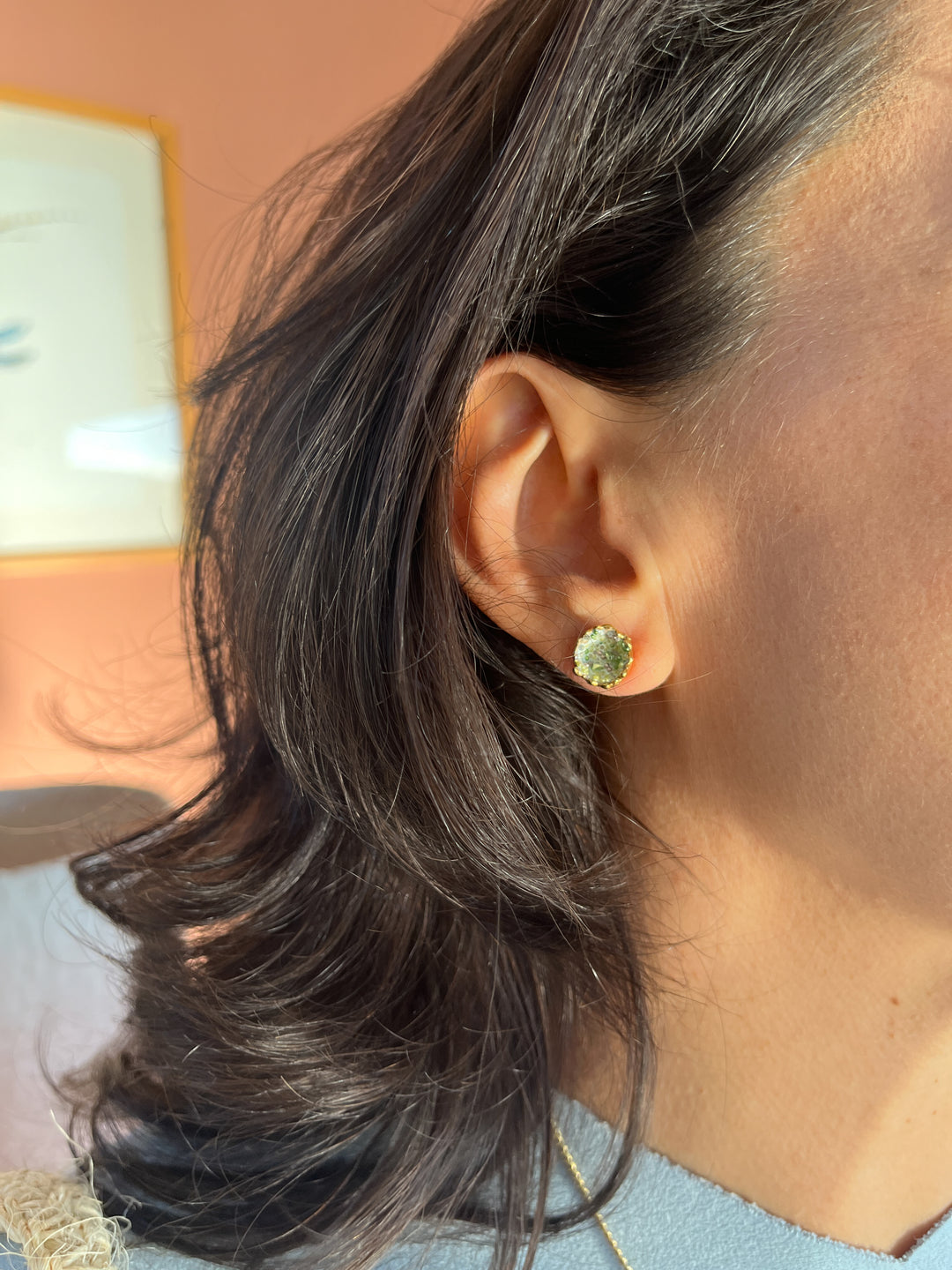 Close-up side view of a woman’s ear wearing a  Coca M stud earring, hand-painted in sage with glass granulation on a gold-plated brass setting, her hair tucked behind her ear against a neutral background.