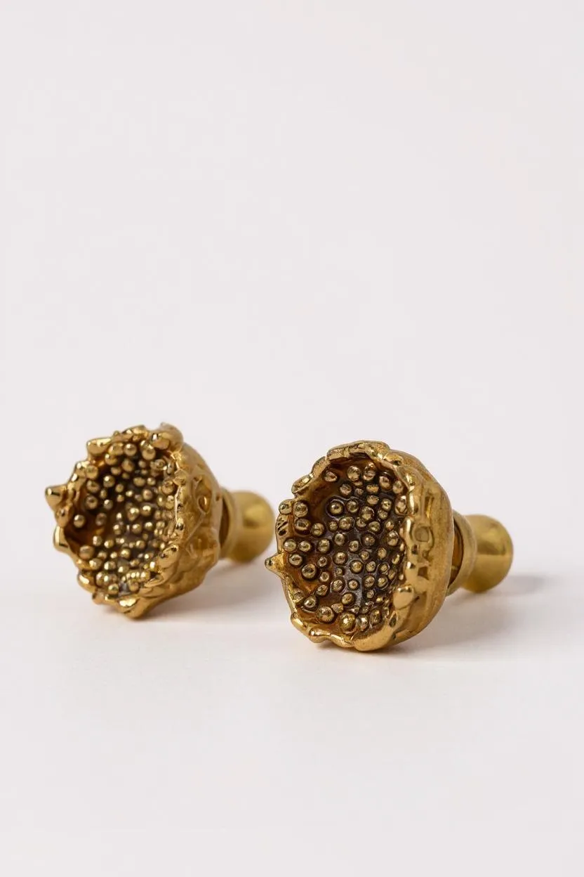 Close-up of two Topos Coca M stud earrings in gold-plated bronze featuring detailed glass granulation texture, displayed on a pale neutral background.