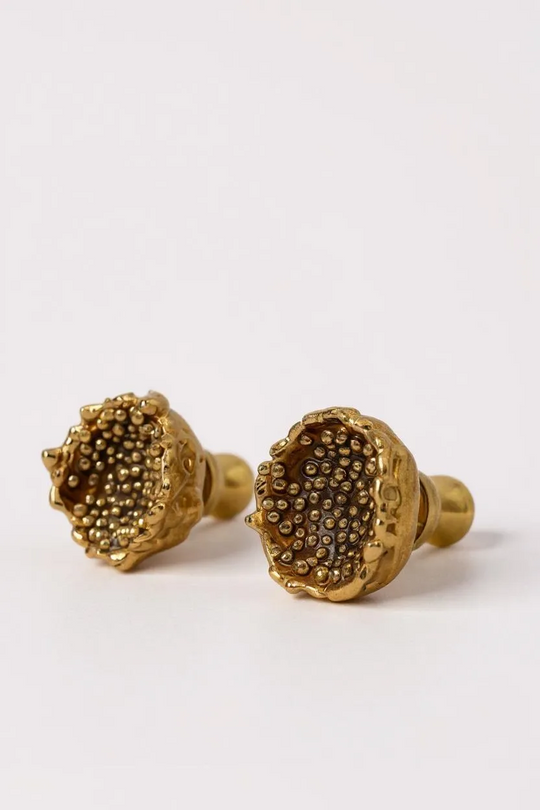 Close-up of two Topos Coca M stud earrings in gold-plated bronze featuring detailed glass granulation texture, displayed on a pale neutral background.