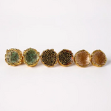 Flat lay of six Topos Coca M stud earrings on a white background, showing all three hand-painted variants—two in sage-green with glass granulation, two in rust with glass granulation, and two in sunset paint with turquoise granulation—each set in gold-plated bronze.