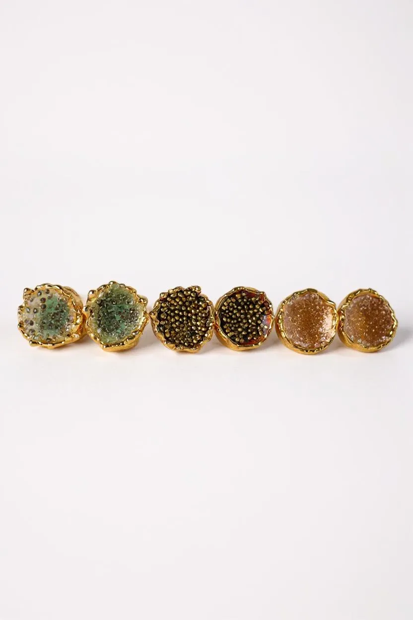 Flat lay of six Topos Coca M stud earrings on a white background, showing all three hand-painted variants—two in sage-green with glass granulation, two in rust with glass granulation, and two in sunset paint with turquoise granulation—each set in gold-plated bronze.