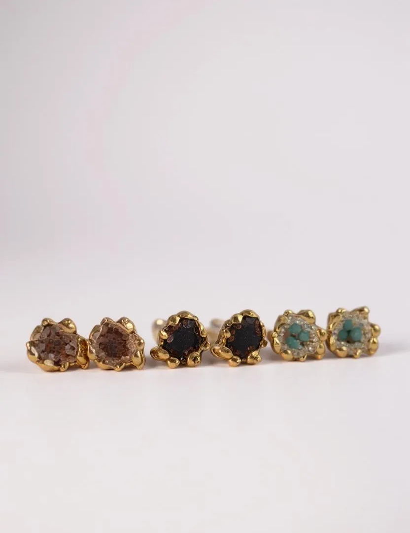 Six Topos Coca S stud earrings arranged in a horizontal line on a pale background, each featuring a scalloped gold-tone setting filled with glass granulation and painted finishes in shades of brown, black, green, turquoise, and more