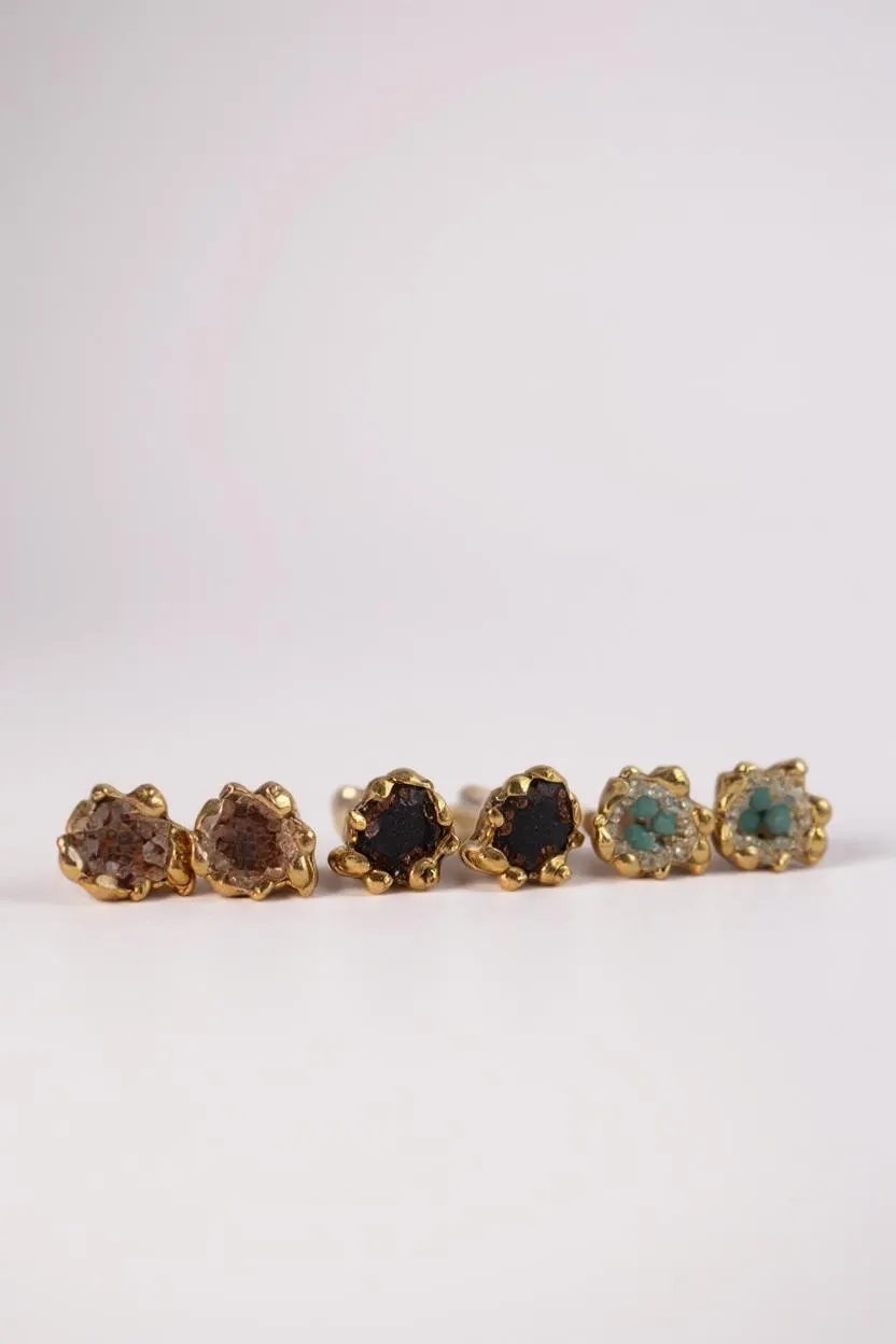 Six Topos Coca S stud earrings arranged in a horizontal line on a pale background, each featuring a scalloped gold-tone setting filled with glass granulation and painted finishes in shades of brown, black, green, turquoise, and more