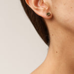 Close-up side view of a woman’s ear wearing a single Topos Coca S stud earring, showcasing its scalloped gold-tone setting filled with green glass granulation and paint.