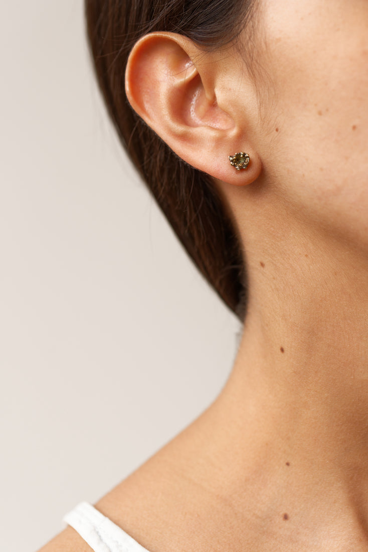 Close-up side view of a woman’s ear wearing a single Topos Coca S stud earring, showcasing its scalloped gold-tone setting filled with green glass granulation and paint.