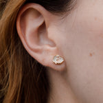 Close-up side view of a woman’s ear wearing a Topos Cucu stud earring hand-painted in rose-pink paint with glass granulation on a gold-plated brass setting, hair tucked behind ear, against a neutral background.