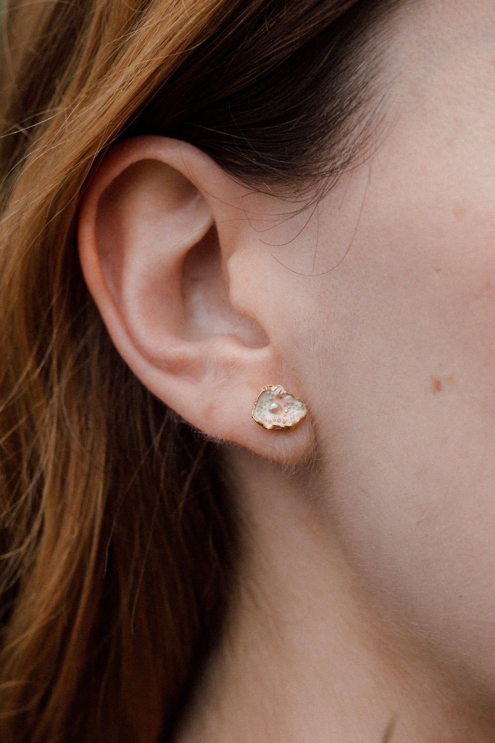 Close-up side view of a woman’s ear wearing a Topos Cucu stud earring hand-painted in rose-pink paint with glass granulation on a gold-plated brass setting, hair tucked behind ear, against a neutral background.