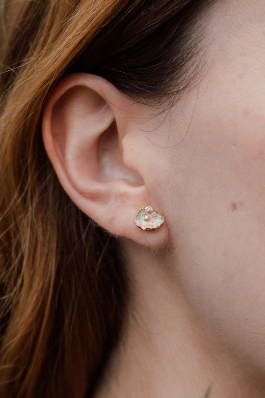 Close-up side view of a woman’s ear wearing a Topos Cucu stud earring hand-painted in rose-pink paint with glass granulation on a gold-plated brass setting, hair tucked behind ear, against a neutral background.