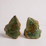 Flat lay of two Topos Cucu stud earrings hand-painted in sage-green paint with glass granulation on gold-plated brass, displayed on a white background.