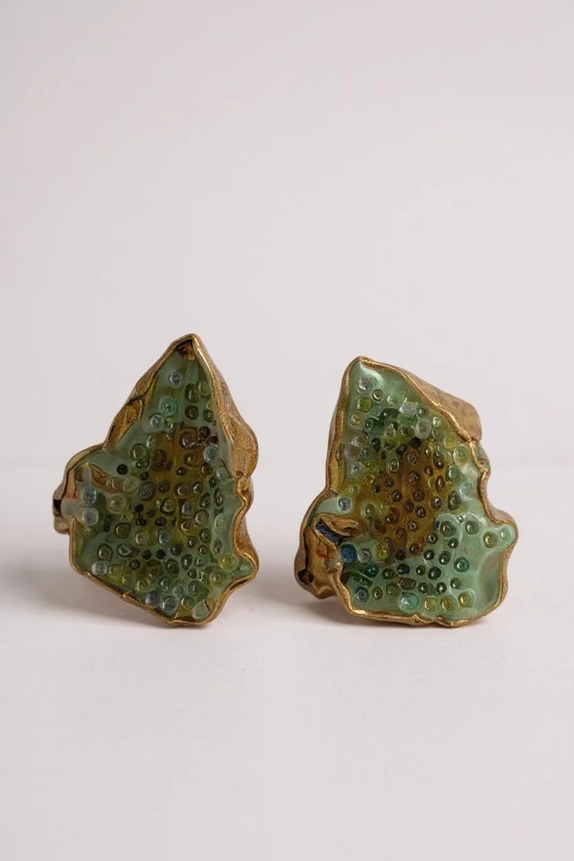 Flat lay of two Topos Cucu stud earrings hand-painted in sage-green paint with glass granulation on gold-plated brass, displayed on a white background.