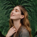 Portrait of a woman wearing a Topos Cucu stud earring hand-painted in pink paint with glass granulation on a gold-plated brass setting, posing against a backdrop of large green leaves.