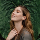 Portrait of a woman wearing a Topos Cucu stud earring hand-painted in pink paint with glass granulation on a gold-plated brass setting, posing against a backdrop of large green leaves.