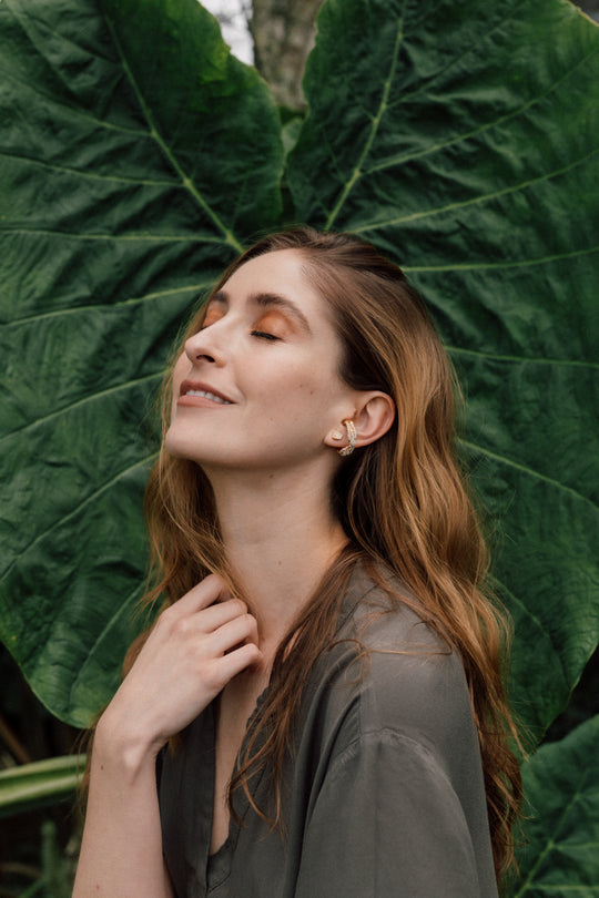 Portrait of a woman wearing a Topos Cucu stud earring hand-painted in pink paint with glass granulation on a gold-plated brass setting, posing against a backdrop of large green leaves.