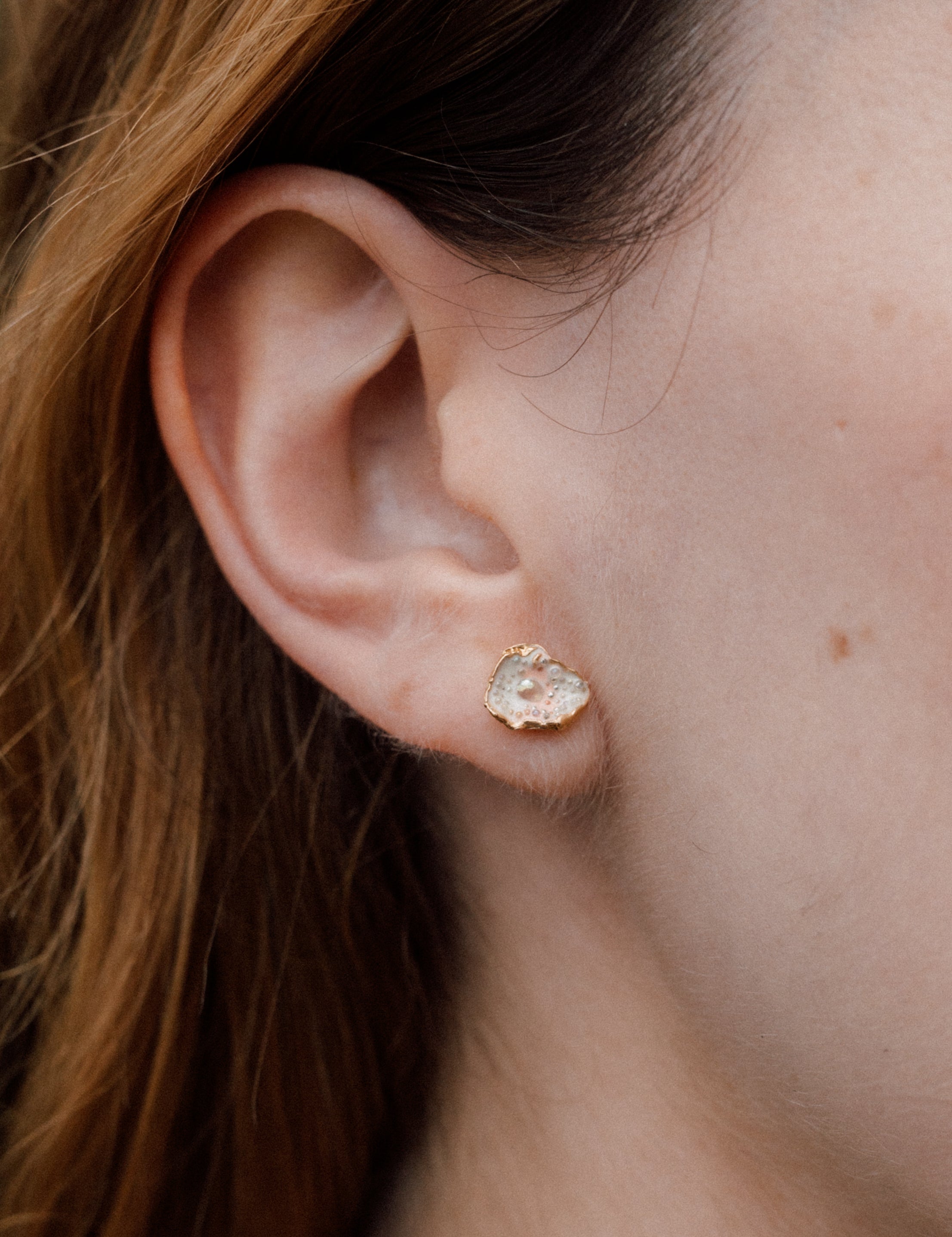 Close-up side view of a woman’s ear wearing a Topos Cucu stud earring hand-painted in rose-pink paint with glass granulation on a gold-plated brass setting, hair tucked behind ear, against a neutral background.