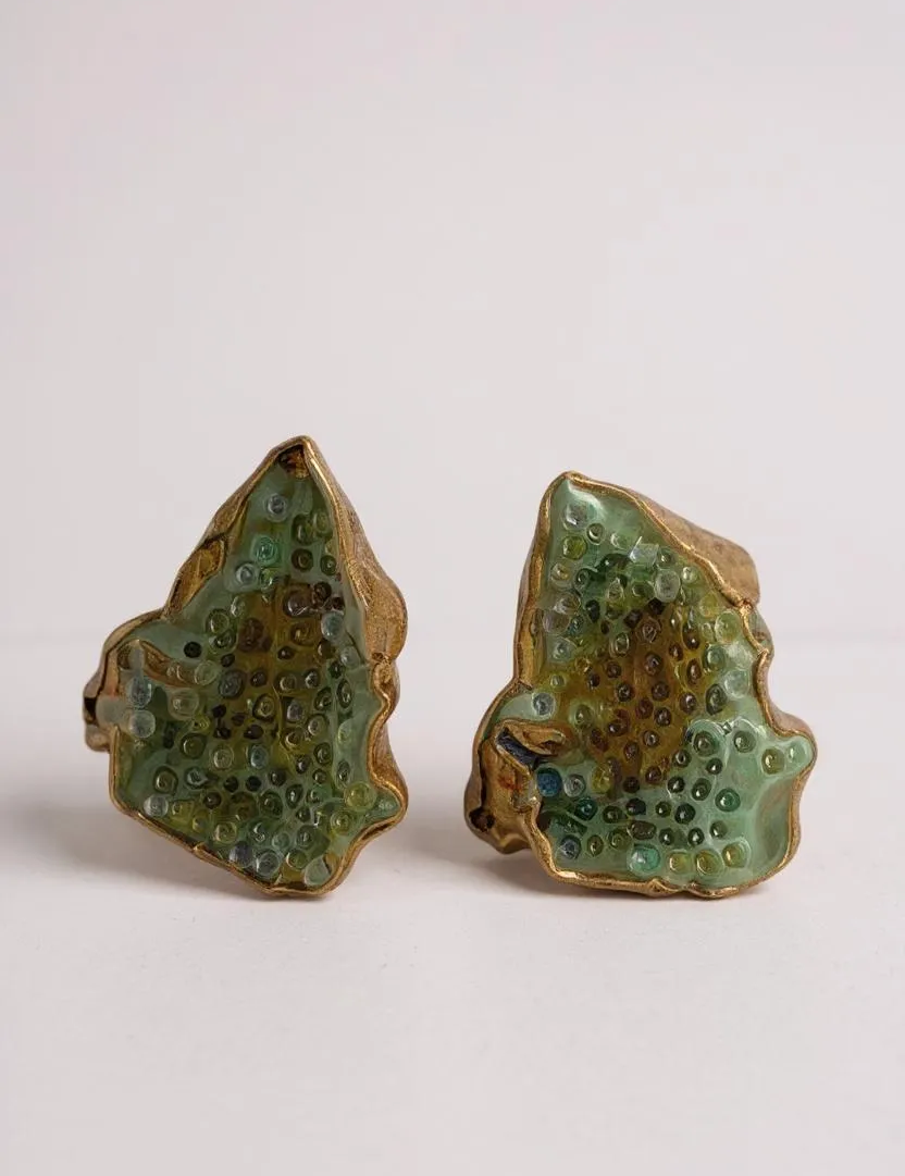 Flat lay of two Topos Cucu stud earrings hand-painted in sage-green paint with glass granulation on gold-plated brass, displayed on a white background.