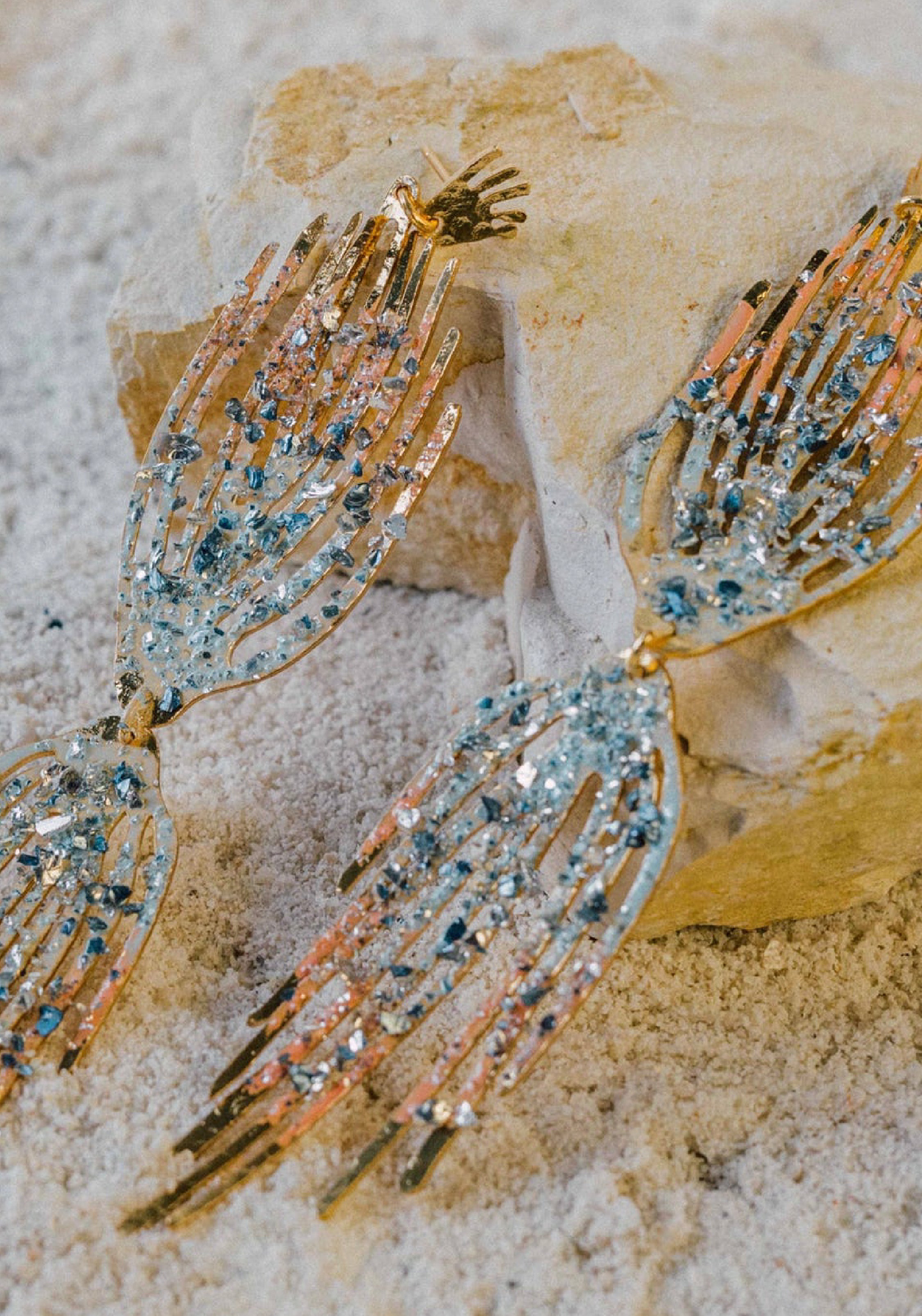 Pair of handcrafted gold earrings shaped like elongated hands, adorned with blue and pink crystals, displayed on sand and stone.