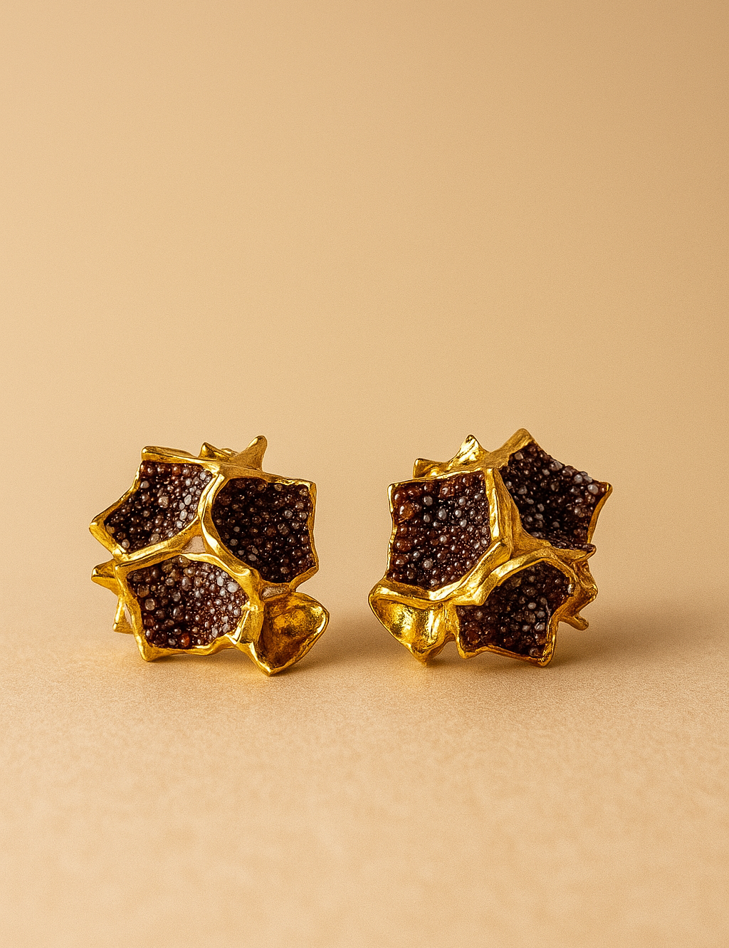 Close-up of a pair of Galaxea stud earrings in rust—gold-plated star-shaped earrings filled with clusters of rust-colored glass granules—on a warm beige background.