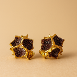  Close-up of a pair of Galaxea stud earrings in rust—gold-plated star-shaped earrings filled with clusters of rust-colored glass granules—on a warm beige background.