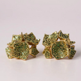 Pair of Galaxea stud earrings in sage—gold-plated floral-shaped earrings with clusters of sage-green glass granules—displayed on a pale pink background.