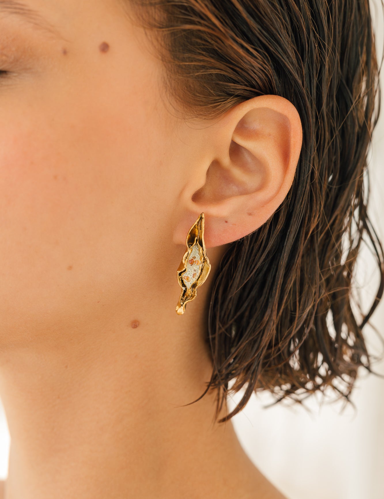 Close-up side view of a woman’s ear wearing the Mustard variant of the Lettuce Leaf Earring, hand-painted mustard-yellow with glass granulation on a gold-plated bronze setting, hair tucked behind ear against a neutral background.