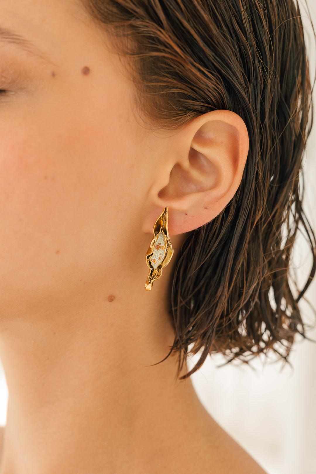 Close-up side view of a woman’s ear wearing the Mustard variant of the Lettuce Leaf Earring, hand-painted mustard-yellow with glass granulation on a gold-plated bronze setting, hair tucked behind ear against a neutral background.